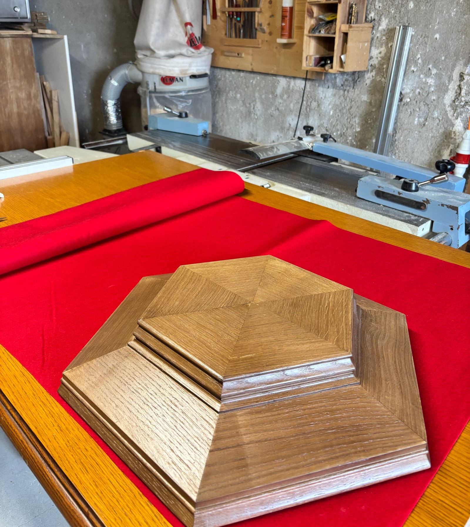 Pièce hexagonale en bois à moulures étagées, posée sur un tissu rouge dans l'atelier.